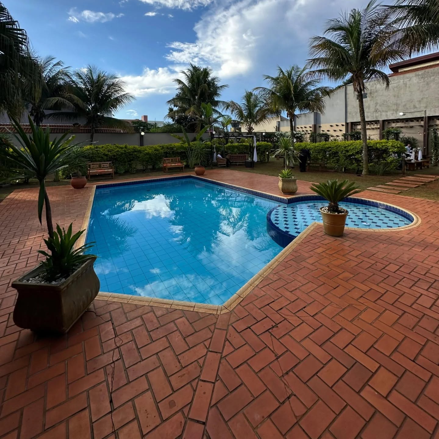Vista ampla da piscina, piso de tijolos, bancos, palmeiras e jardim da casa